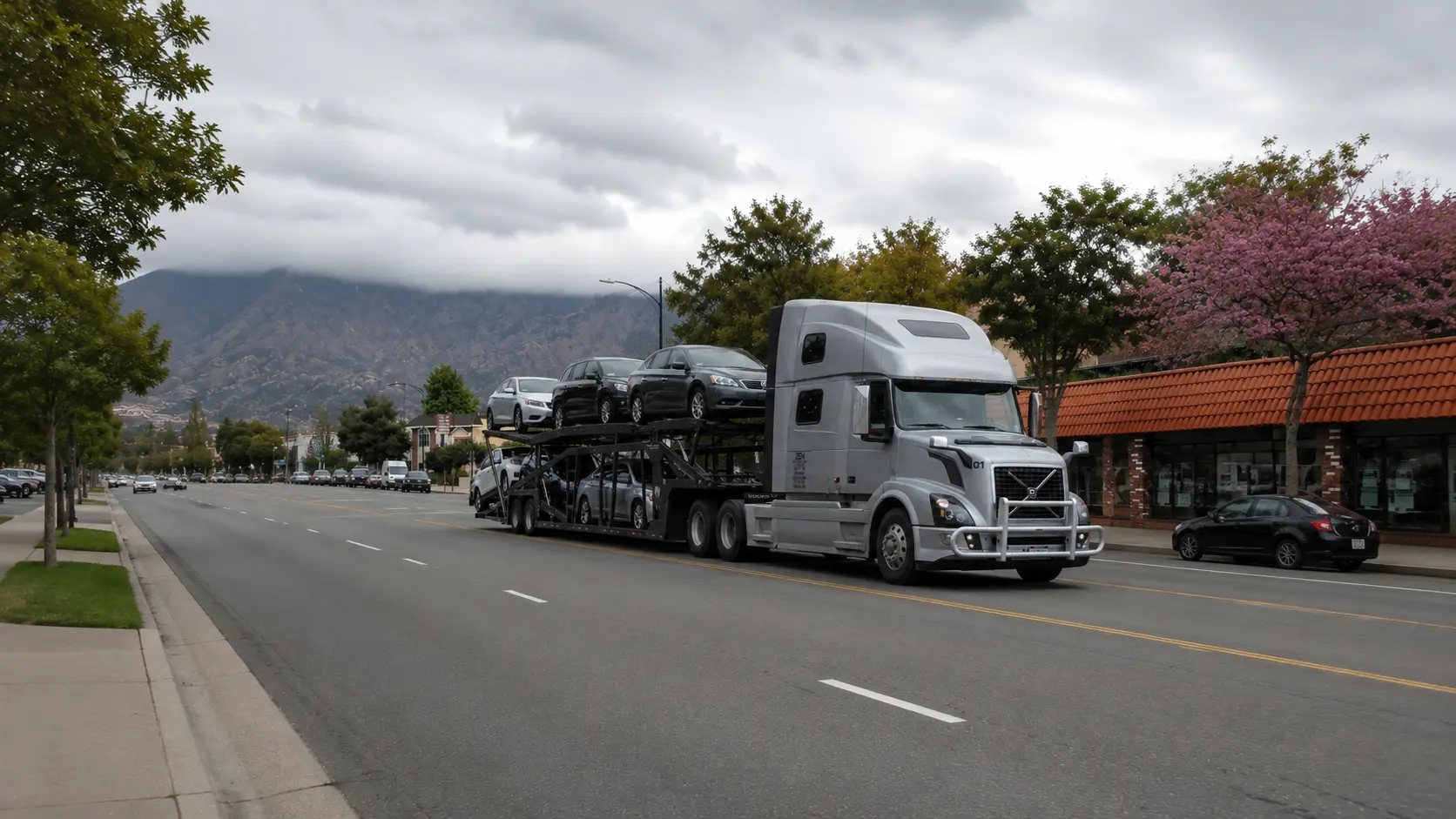 Open car carrier transporting multiple vehicles on a city road with mountains in the background.